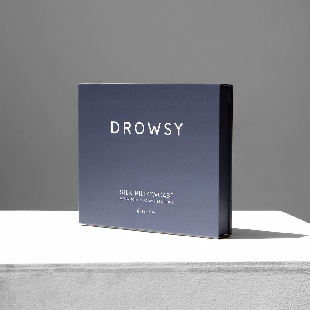 A dark blue rectangular box labeled Drowsy stands on a white surface against a plain gray background, showcasing the Moonlight Shadow Silk Pillowcase—22 Momme, Queen Size—ideal for skincare and hair protection.
