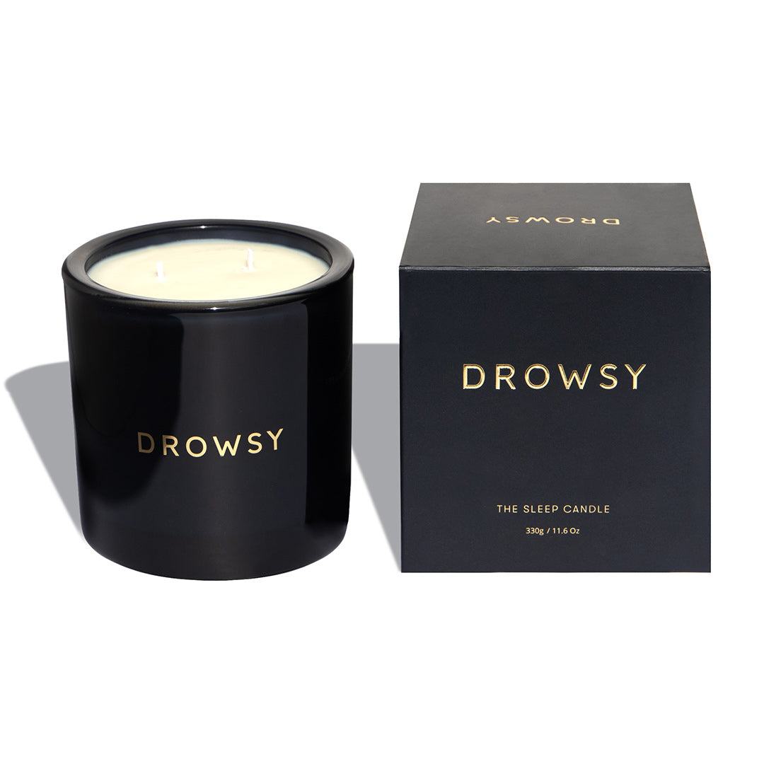 A black candle jar labeled DROWSY sits next to its matching black box, which also displays the word DROWSY and the text THE SLEEP CANDLE 335g / 11.8 oz in gold lettering.