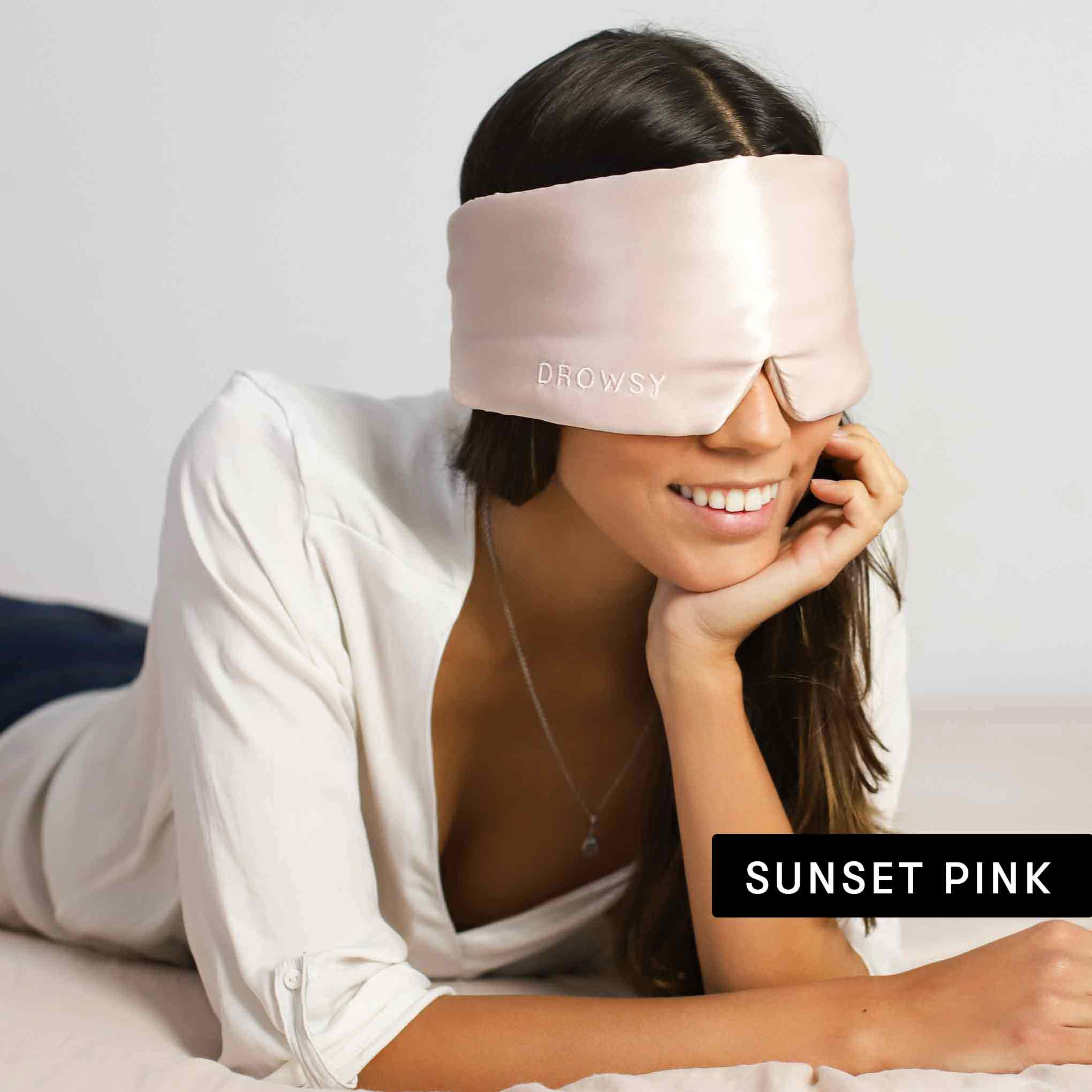 A woman smiles while lying on a bed, wearing a light pink satin sleep mask from the Lovers Pack labeled "DROWSY." She rests her chin on her hand. A text box in the corner reads "SUNSET PINK.