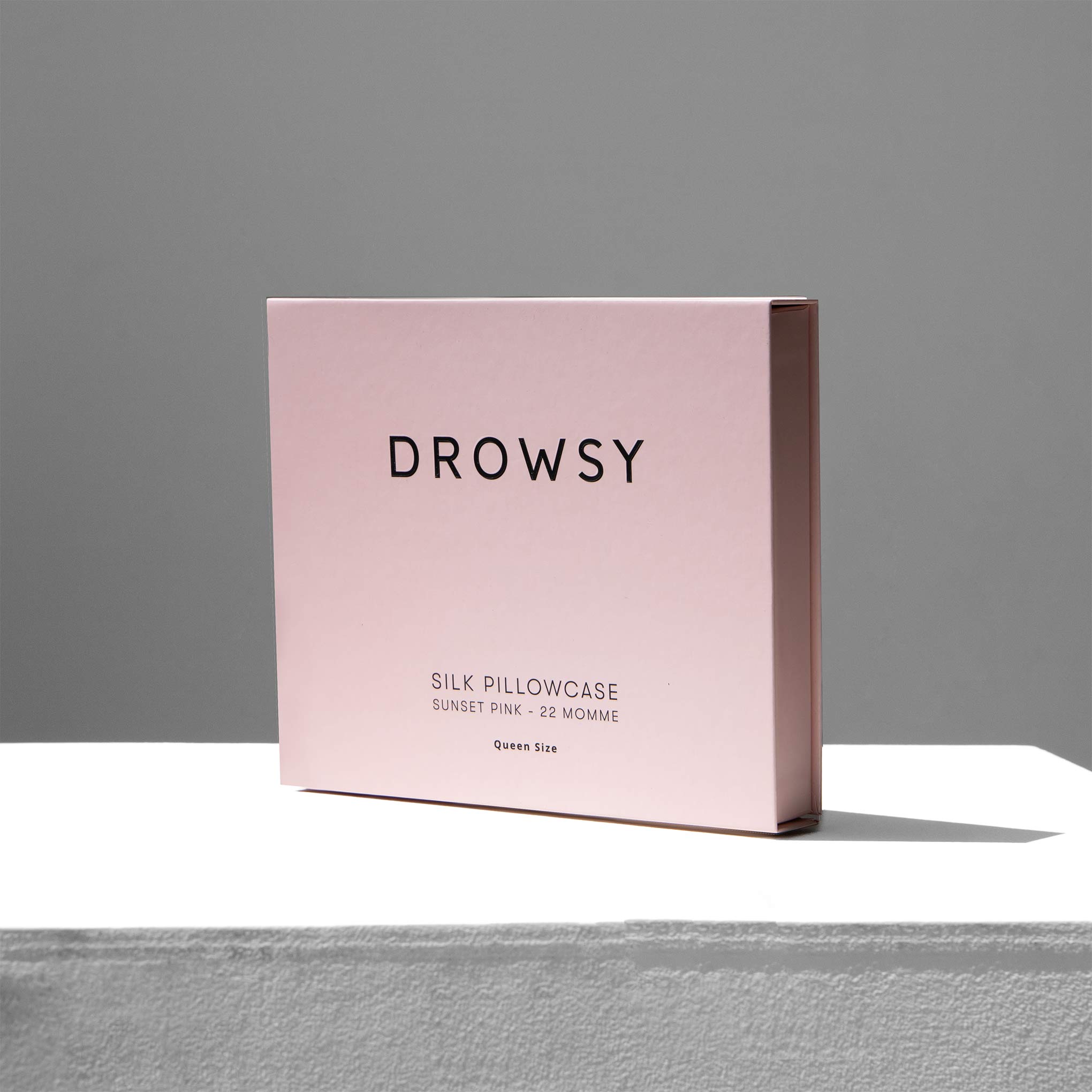 A pink rectangular box labeled DROWSY holds a Sunset Pink Silk Pillowcase. Below, the text reads 22 Momme Queen Size. The box sits on a white surface with a gray background.