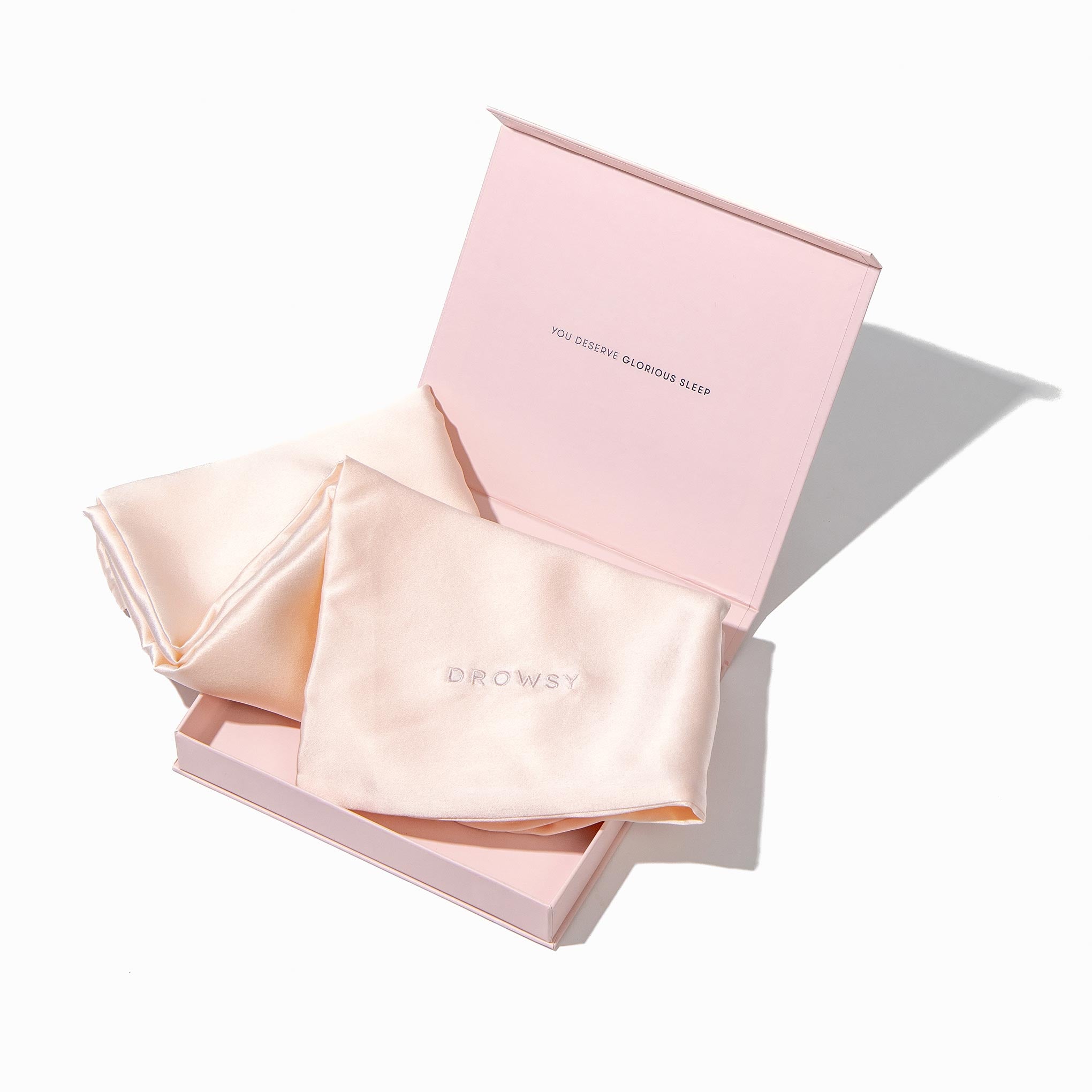 A light pink box with "YOU DESERVE GLAMOROUS SLEEP" inside the lid holds a folded, silky champagne-colored Sunset Pink Silk Pillowcase embroidered with DROWSY. A matching fabric piece beside it is perfect for skincare routines.