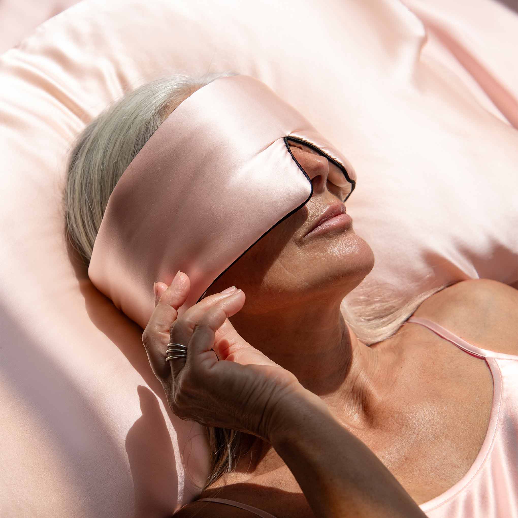 A gray-haired woman wearing a pink satin sleep mask rests peacefully on the Sunset Pink Silk Pillowcase and sheets, gently adjusting her mask while enjoying the hair-protecting benefits as she sleeps.