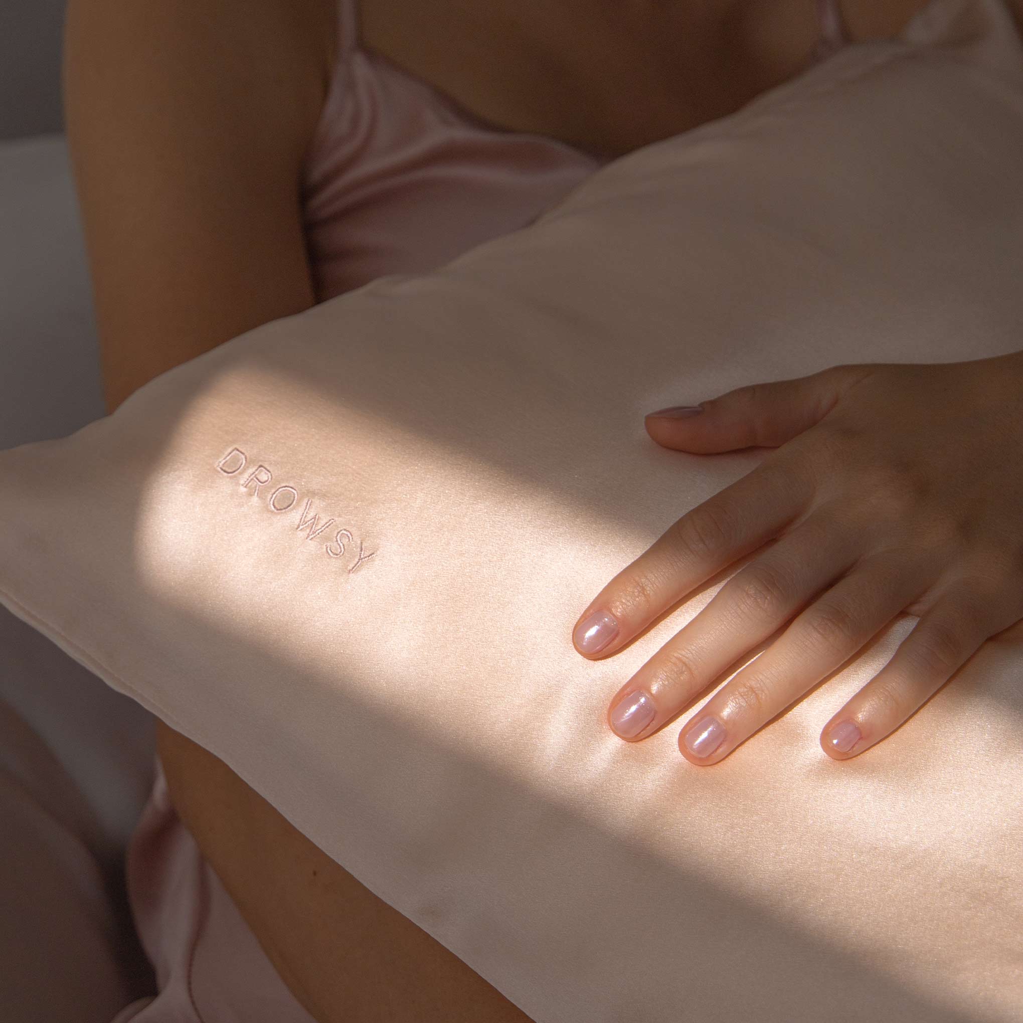 A person in a light pink satin garment rests their hand on the Sunset Pink Silk Pillowcase, embroidered with "DROWSY" and bathed in soft natural light—a luxurious accessory for skincare and hair protection.