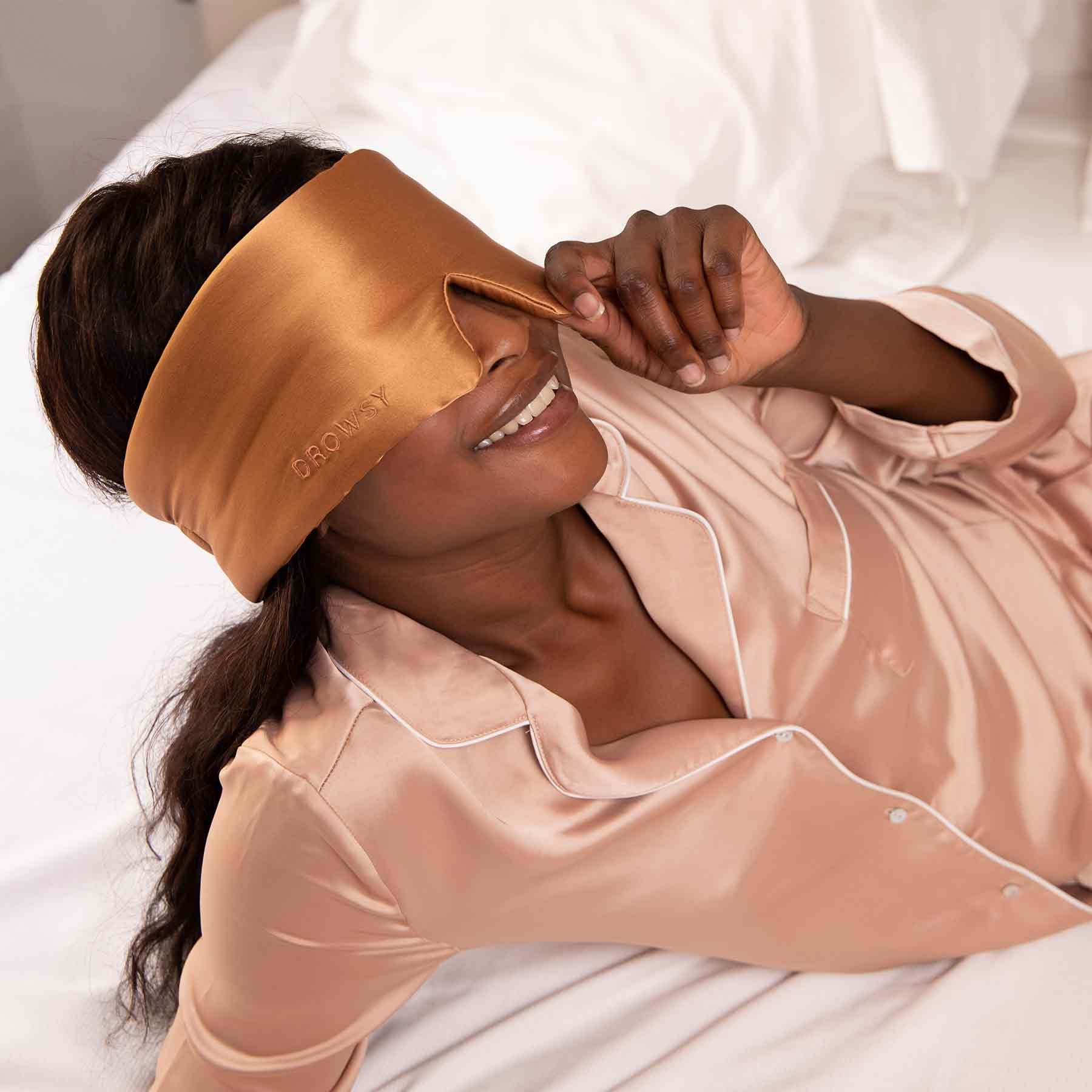 A person in peach silk pajamas lies on a bed while smiling and adjusting a wide, satin eye mask in a matching color. The bedding is white and the mood is relaxed and comfortable.