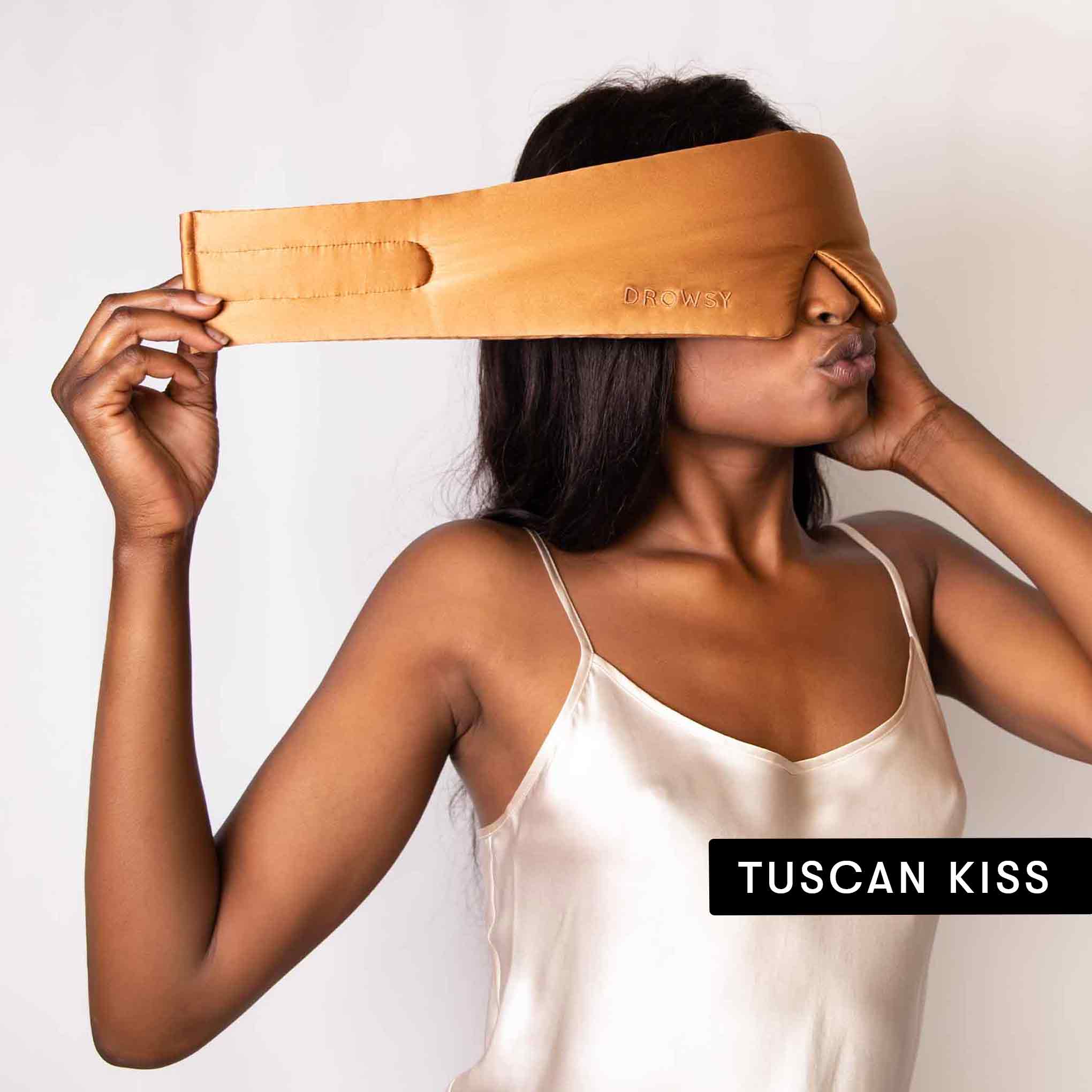 A woman in a cream satin camisole puts on a tan sleep mask labeled DROWSY, with “Lovers Pack” displayed in a black box in the lower right corner.
