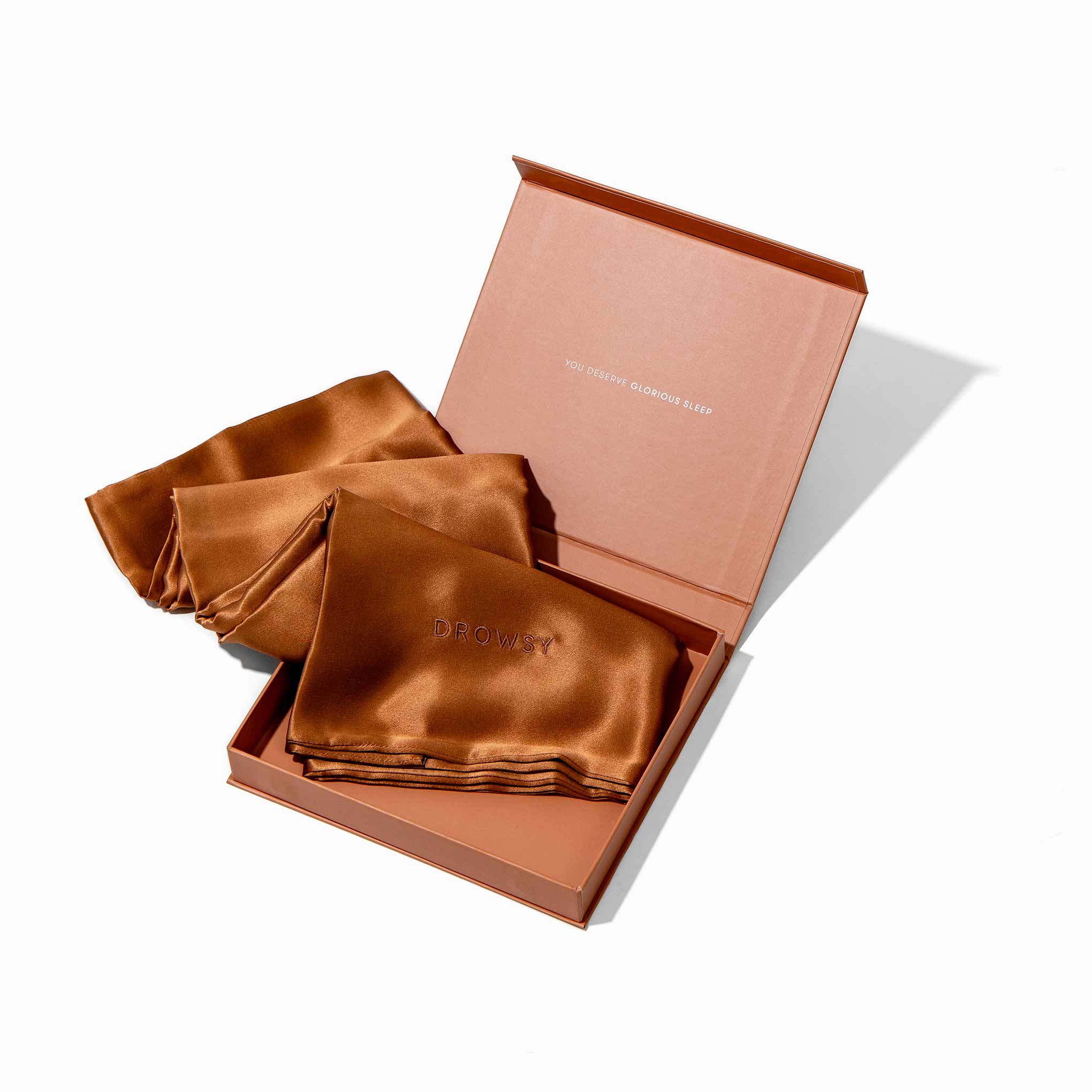 The Pumpkin Spice Silk Pillowcase and matching eye mask, both hair-protecting and essential for skincare, rest inside an elegant open box with text on the lid, displayed against a white background.
