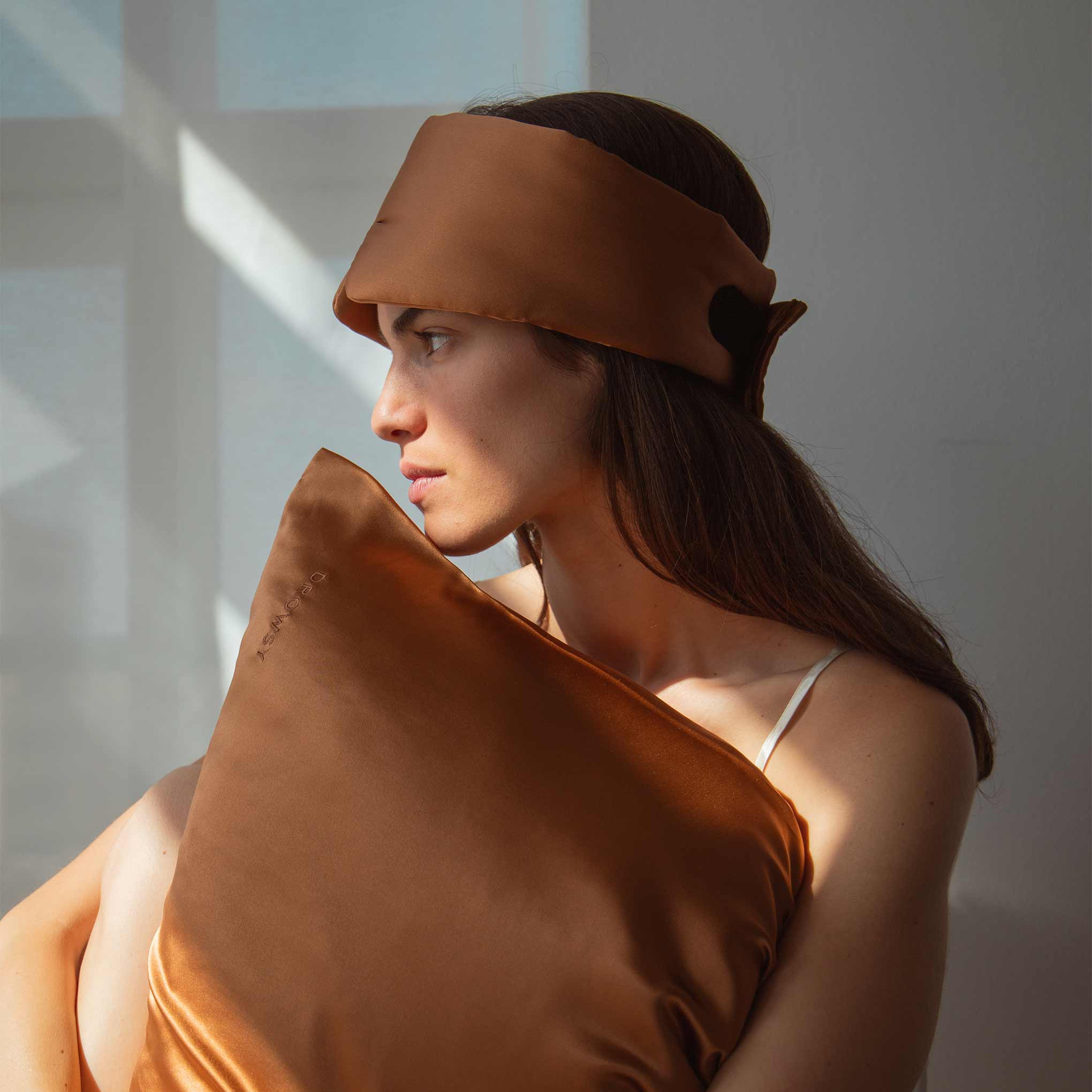 A woman with long brown hair in a white camisole sits by a sunlit window, wearing a brown satin sleep mask and holding the Pumpkin Spice Silk Pillowcase—an essential for hair protection, skincare, and beauty routines.
