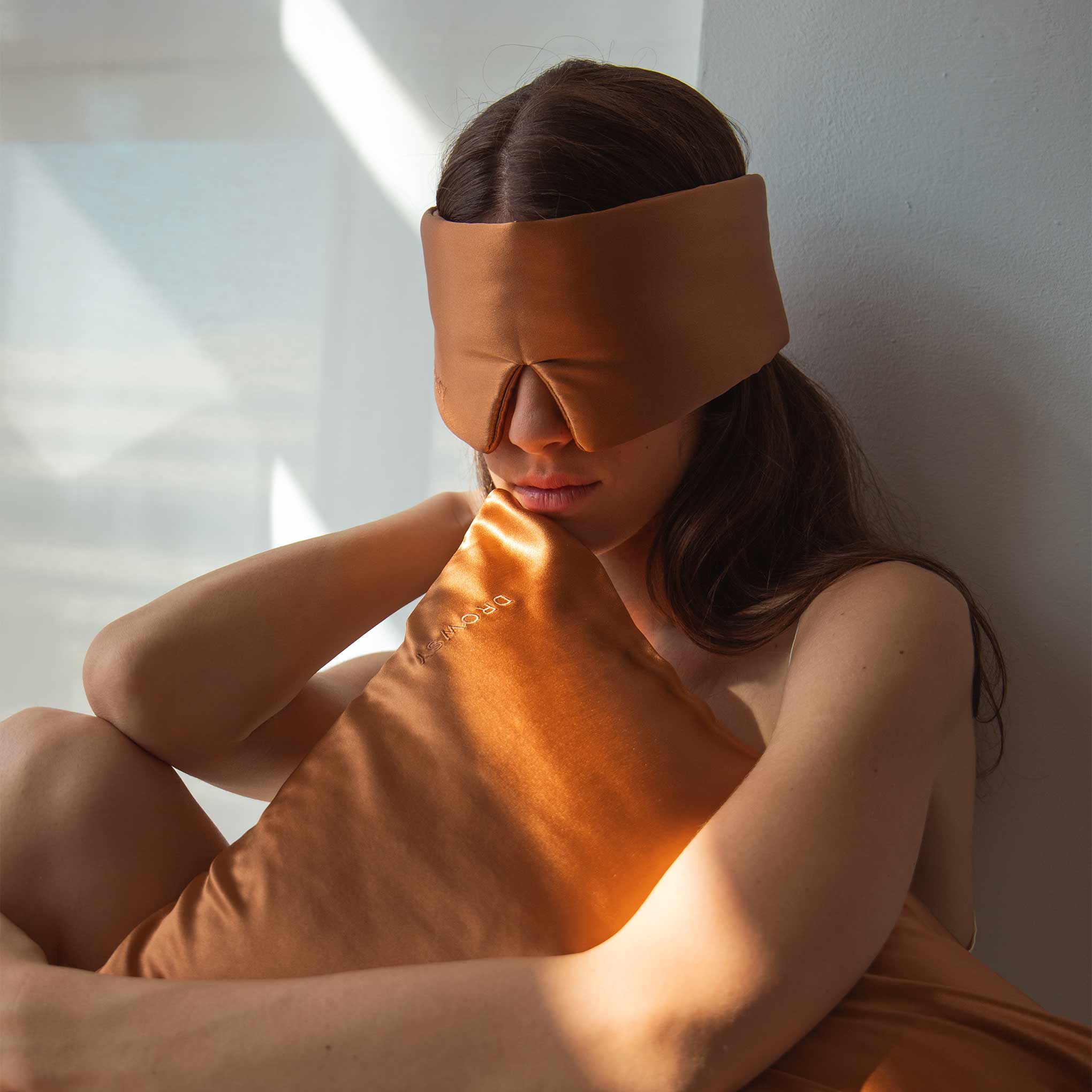 A woman sits against a wall, wearing a brown sleep mask and holding the Pumpkin Spice Silk Pillowcase—an essential for skincare and beauty—as sunlight streams in, casting soft light and shadows around her.