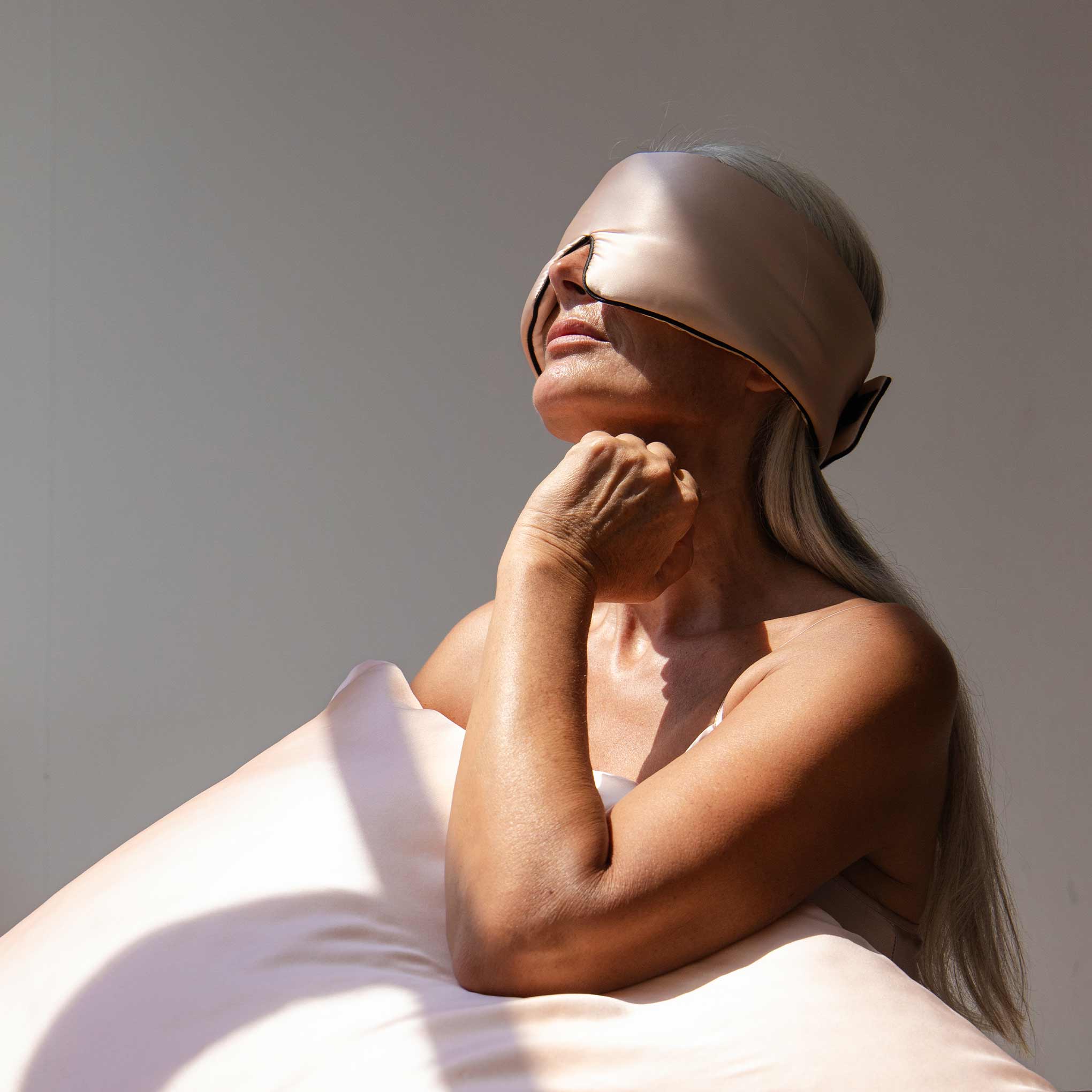 A woman with long gray hair wears the Sunset Pink Pipe Dreams Edition silk sleep mask and gently rests her chin on her hand while holding a matching satin pillow, bathed in soft sunlight against a plain background.