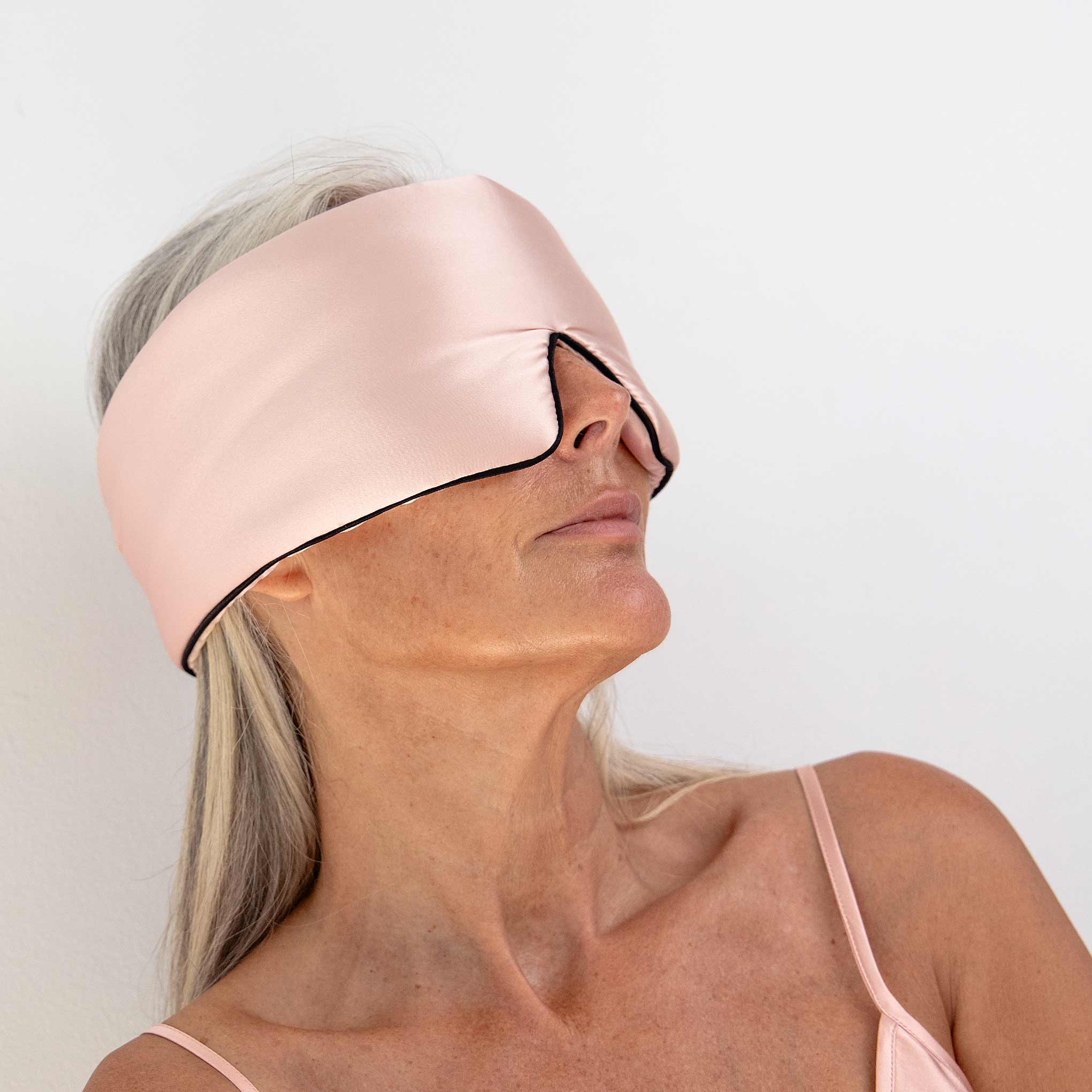 A woman with long gray hair wears the Sunset Pink Pipe Dreams Edition wide silk sleep mask, covering her eyes and part of her nose. She is dressed in a light pink spaghetti strap top and faces slightly upward against a plain white background.