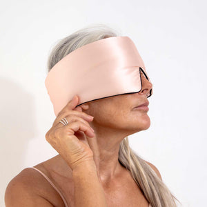 A woman with long gray hair, shown in profile against a plain white background, wears the Sunset Pink Pipe Dreams Edition silk sleep mask and gently touches its edge with her right hand.