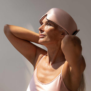 A woman with light skin and long gray hair wears the Sunset Pink Pipe Dreams Edition sleeveless top and matching padded silk sleep mask as a headband, looking upward with her hands behind her head in soft natural light.