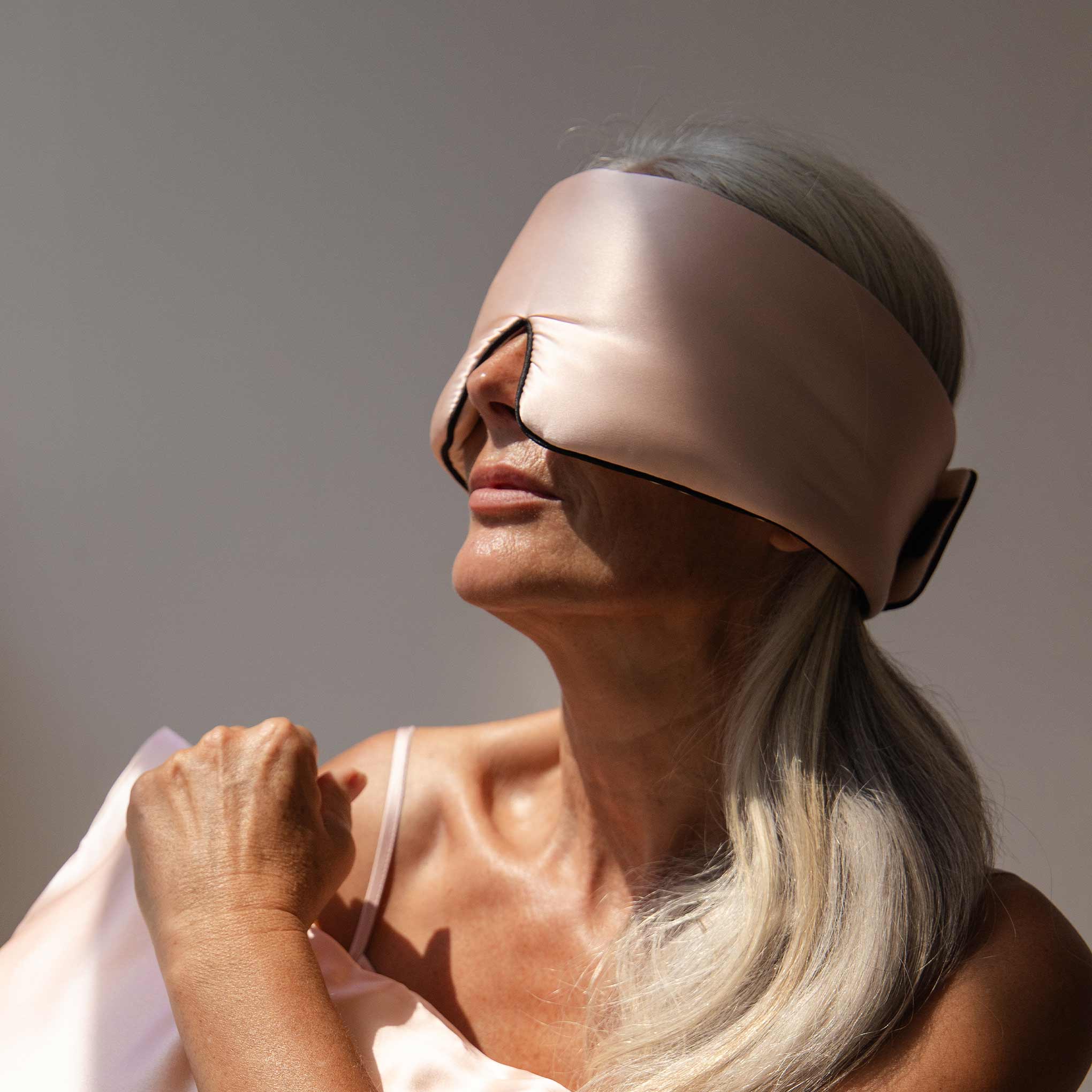 A woman with long gray hair wears the Sunset Pink Pipe Dreams Edition, a large adjustable silk sleep mask covering her eyes and part of her face, bathed in soft light and holding matching satin fabric for nighttime comfort.