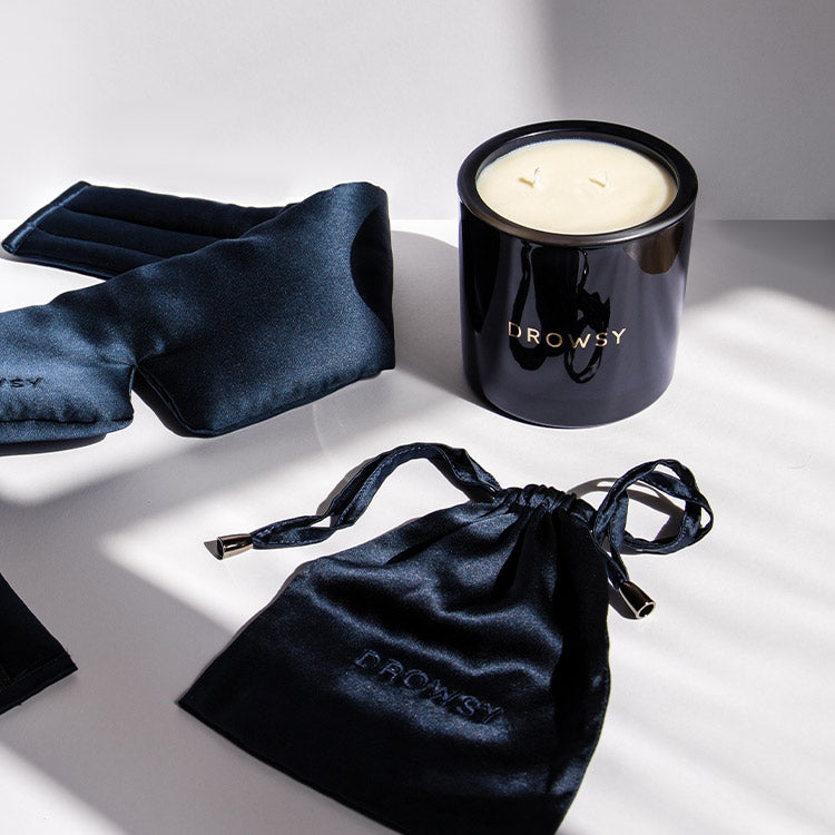 A black candle labeled Drowsy, a black silk sleep mask, and a matching black drawstring pouch are arranged on a white surface in bright natural light.