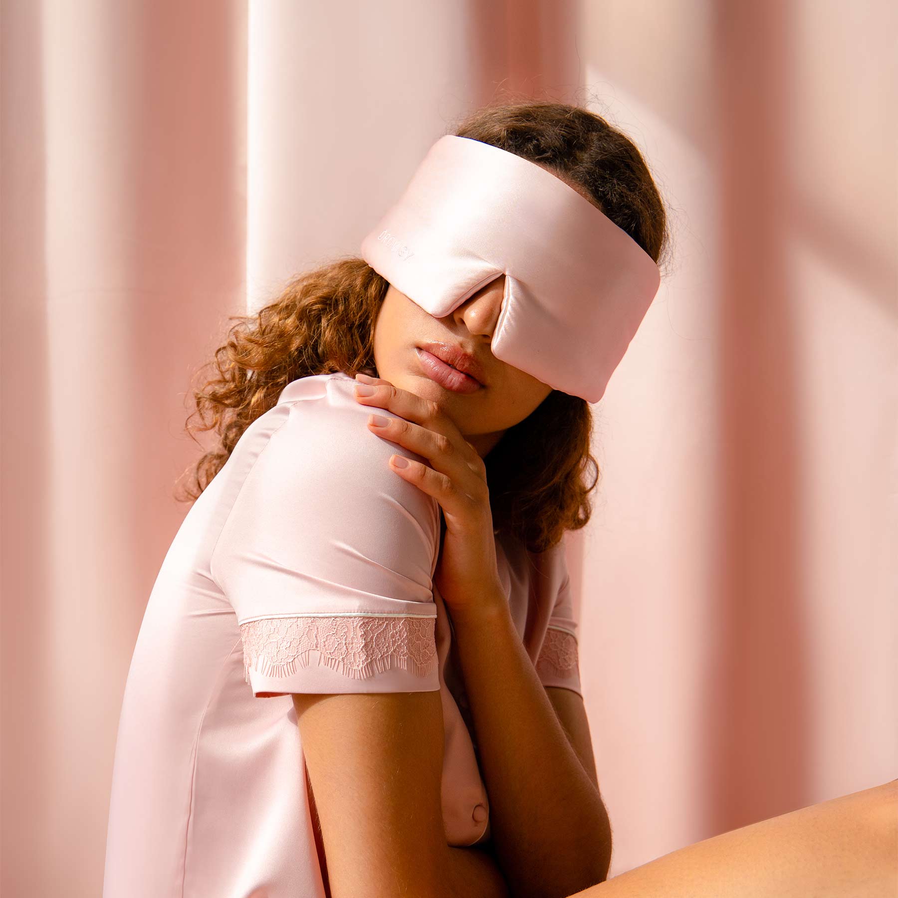 Wearing Sunset Pink silk eye mask, a person with curly hair and pale pink pajamas sits against a pink background, gently resting one hand on their shoulder as they embrace restful sleep and protect against dark circles.