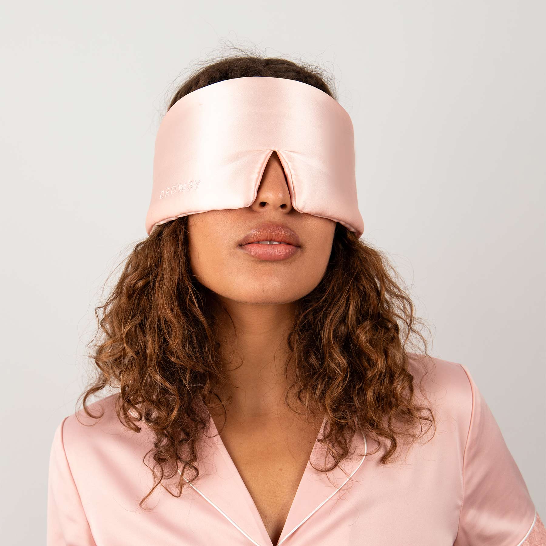 A woman with curly hair wears the Sunset Pink silk eye mask and matching satin pajamas, standing against a white background. The mask covers her eyes, offering beauty sleep and helping prevent dark circles.
