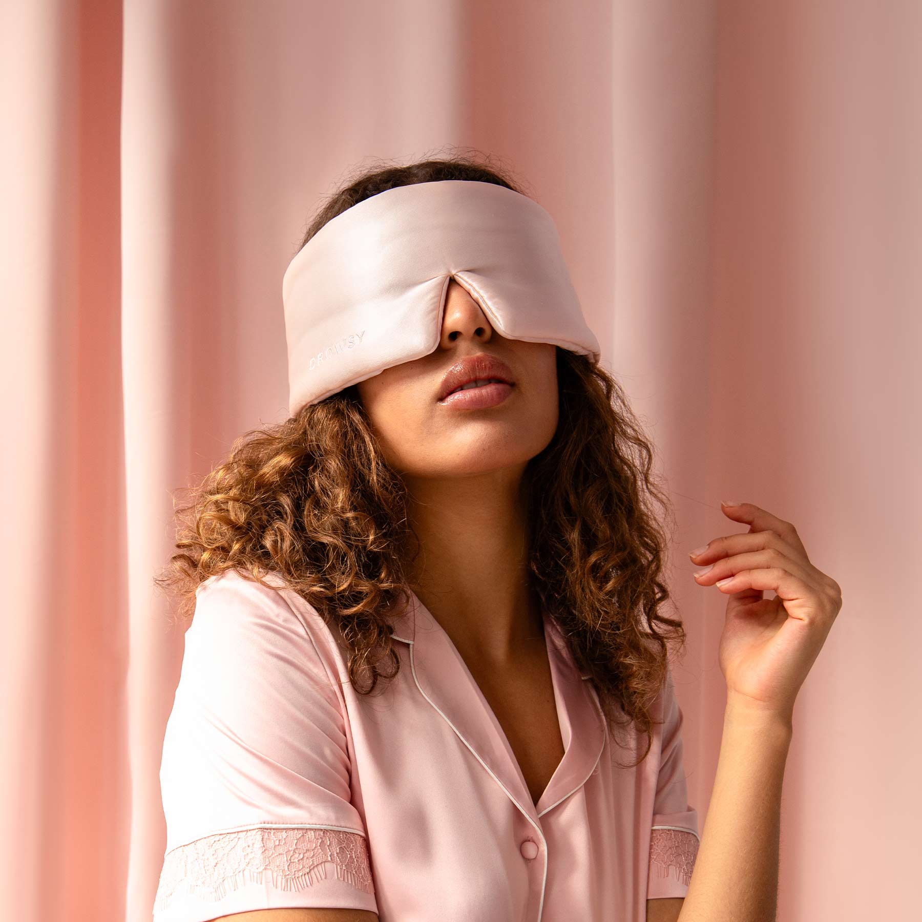 A woman with curly hair, wearing Sunset Pink pajamas and a silk eye mask, poses against a matching background—raising her hand by her face, ready for restful beauty sleep and to help prevent dark circles.