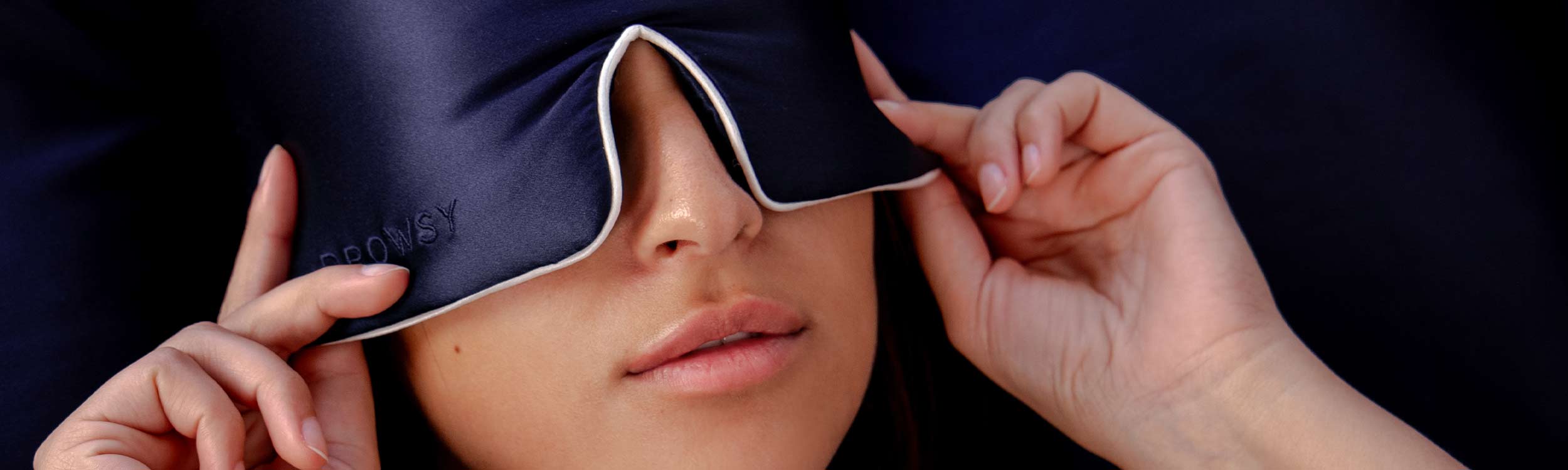 A person lies down, wearing a dark blue silky sleep mask that partially covers their closed eyes. Both hands gently adjust the edges of the mask.