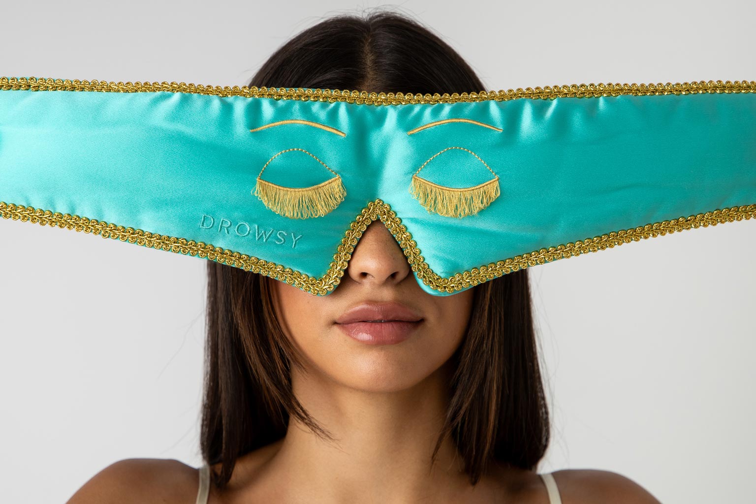 A woman with straight dark hair wears a large turquoise and gold sleep mask embroidered with closed eyes and fringe detailing. The mask covers her eyes and forehead.