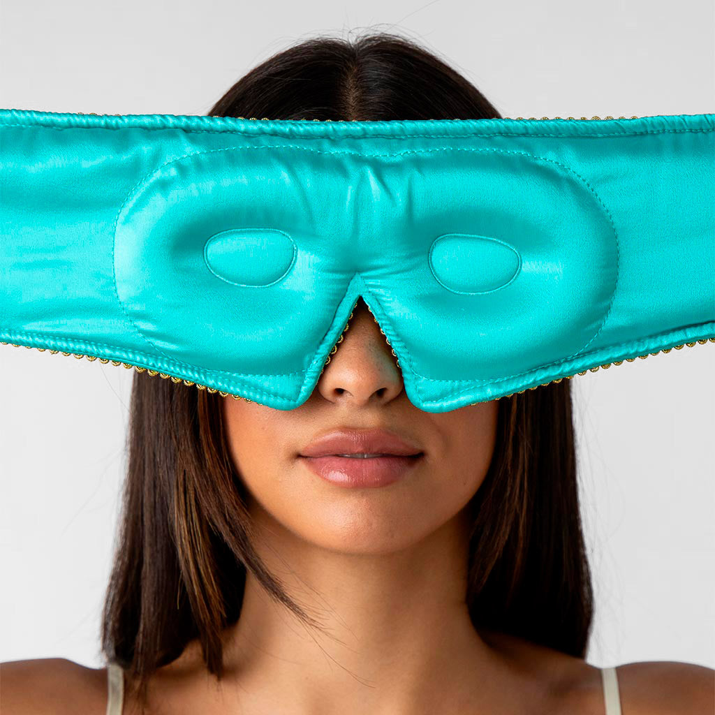 A woman with straight, dark hair wears a turquoise eye mask over her eyes and nose, leaving her mouth and chin visible, against a plain light background.