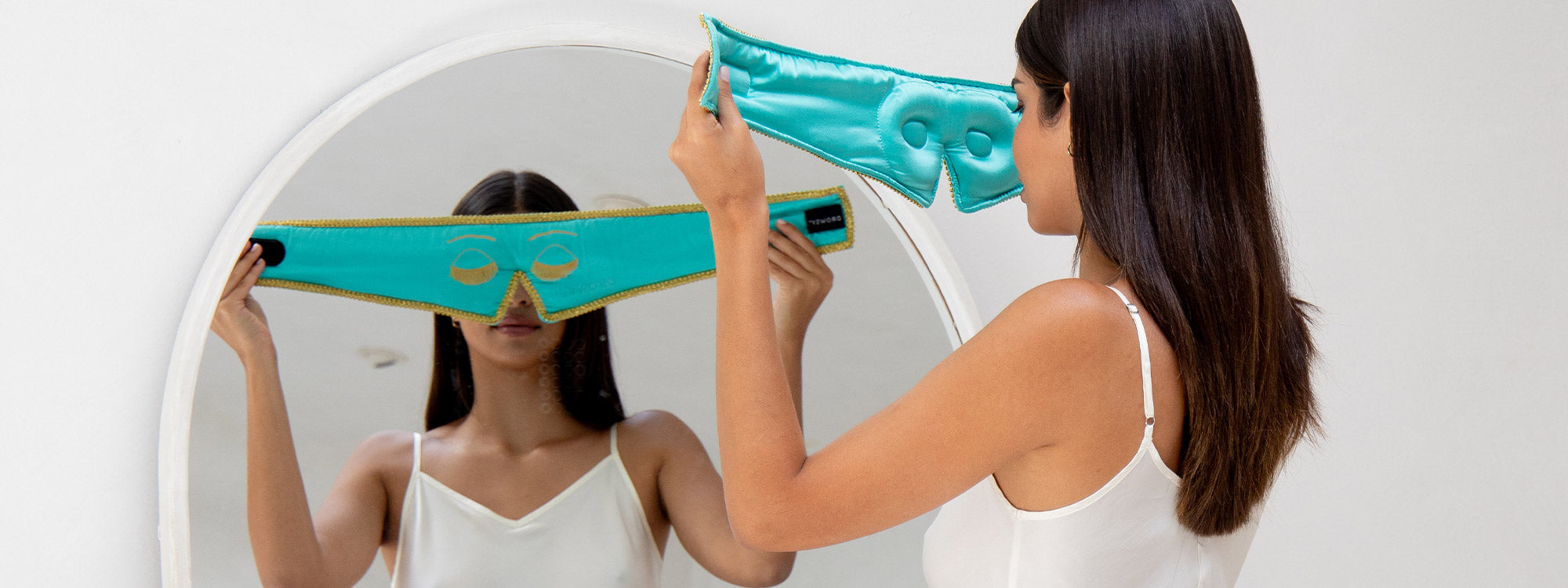 A woman in a white camisole stands in front of a mirror, holding up a turquoise eye mask with cutouts, preparing to put it on her face.