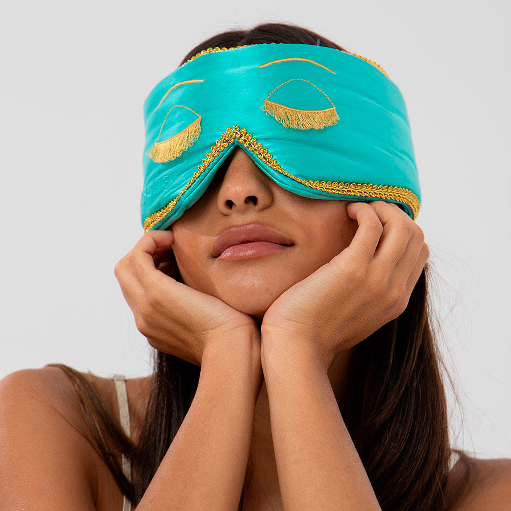 A woman rests her face in her hands while wearing a turquoise sleep mask decorated with golden embroidered closed eyes and lashes.