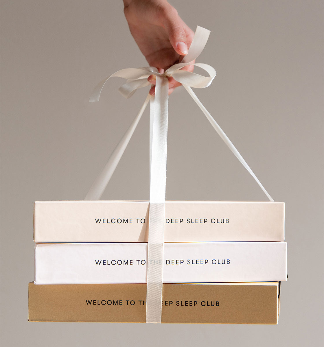 A hand holds three stacked gift boxes tied with a white ribbon. The boxes are beige, white, and gold, each labeled Welcome to the Deep Sleep Club. The background is neutral and plain.