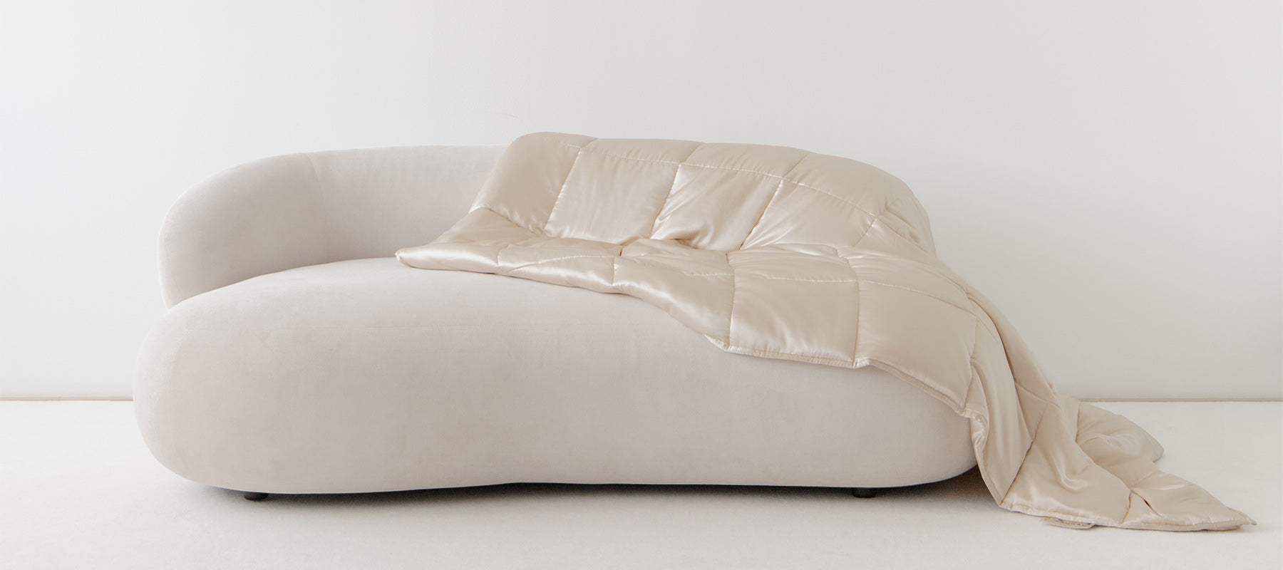 A minimalist, off-white curved sofa with a beige, satin-like quilt draped casually over one side, set against a plain white wall and floor.