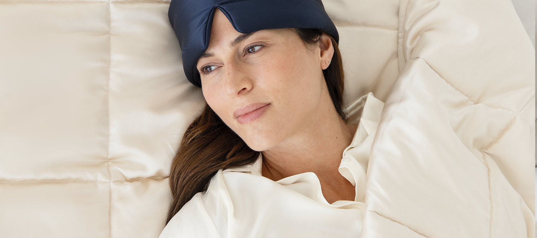 A woman wearing a dark sleep mask on her forehead lies in bed under a cream-colored blanket, looking relaxed and thoughtful.