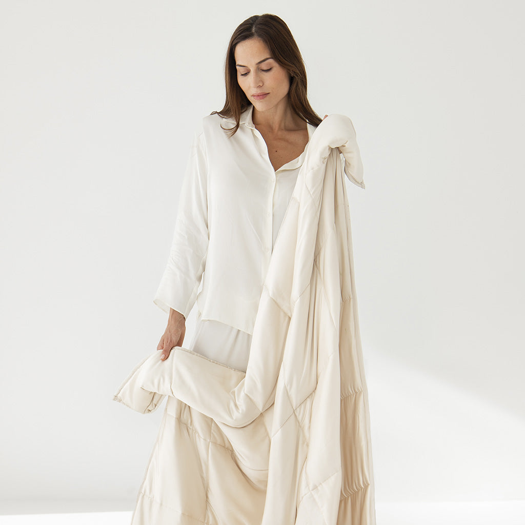 A woman wearing a loose white shirt holds and drapes a large, cream-colored quilt or blanket over her arm while standing against a plain white background.