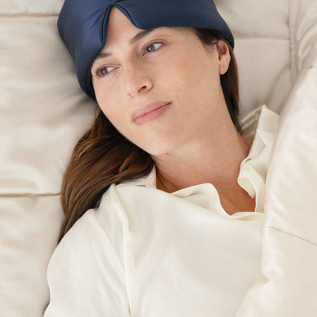 A woman with long brown hair relaxes in bed under a Dusty Gold Silk Weighted Blanket, wearing a white top and a navy blue sleep mask pushed up on her forehead, enjoying deep relaxation.