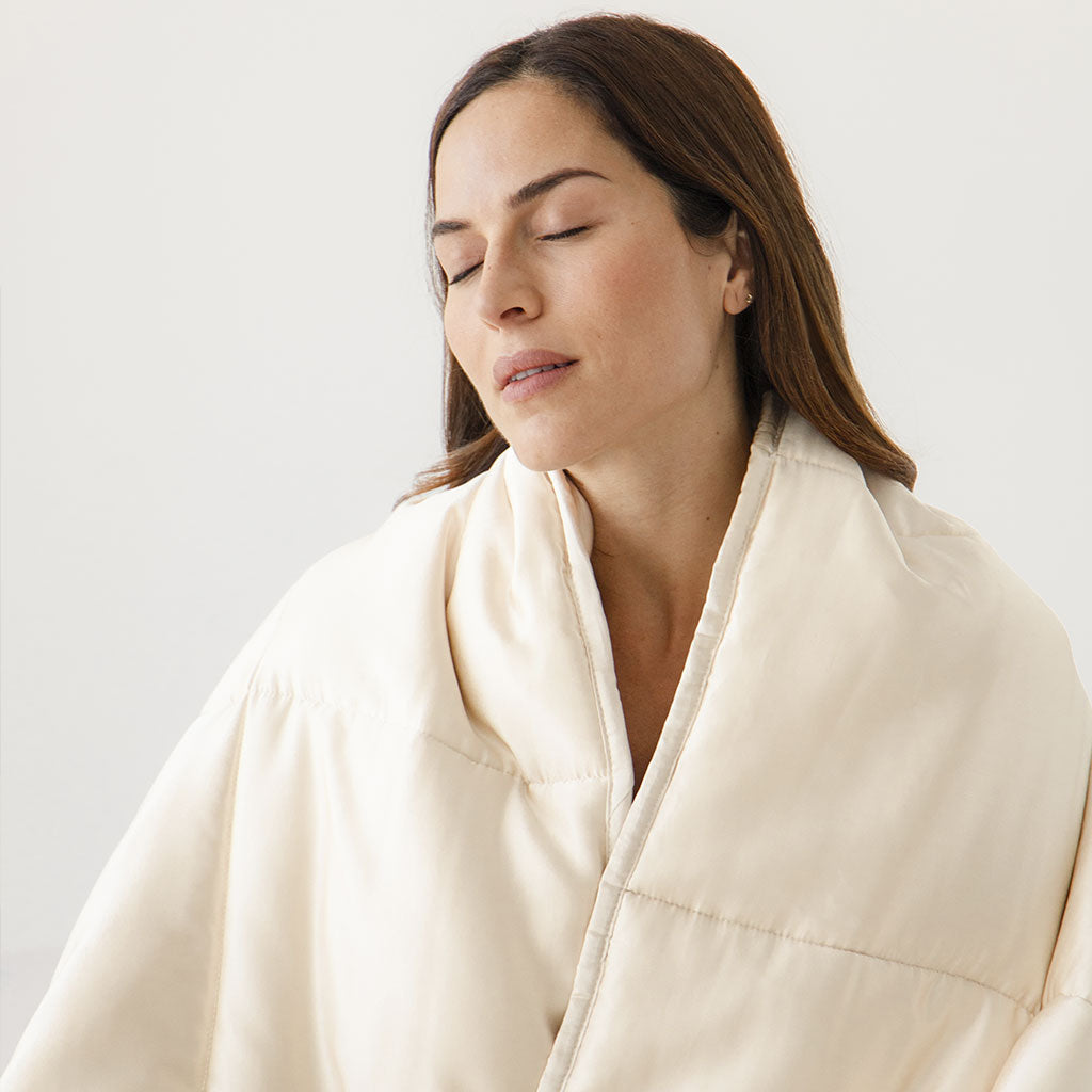 A woman with long brown hair and closed eyes is wrapped in a soft, cream-colored blanket, appearing relaxed and content against a plain light background.