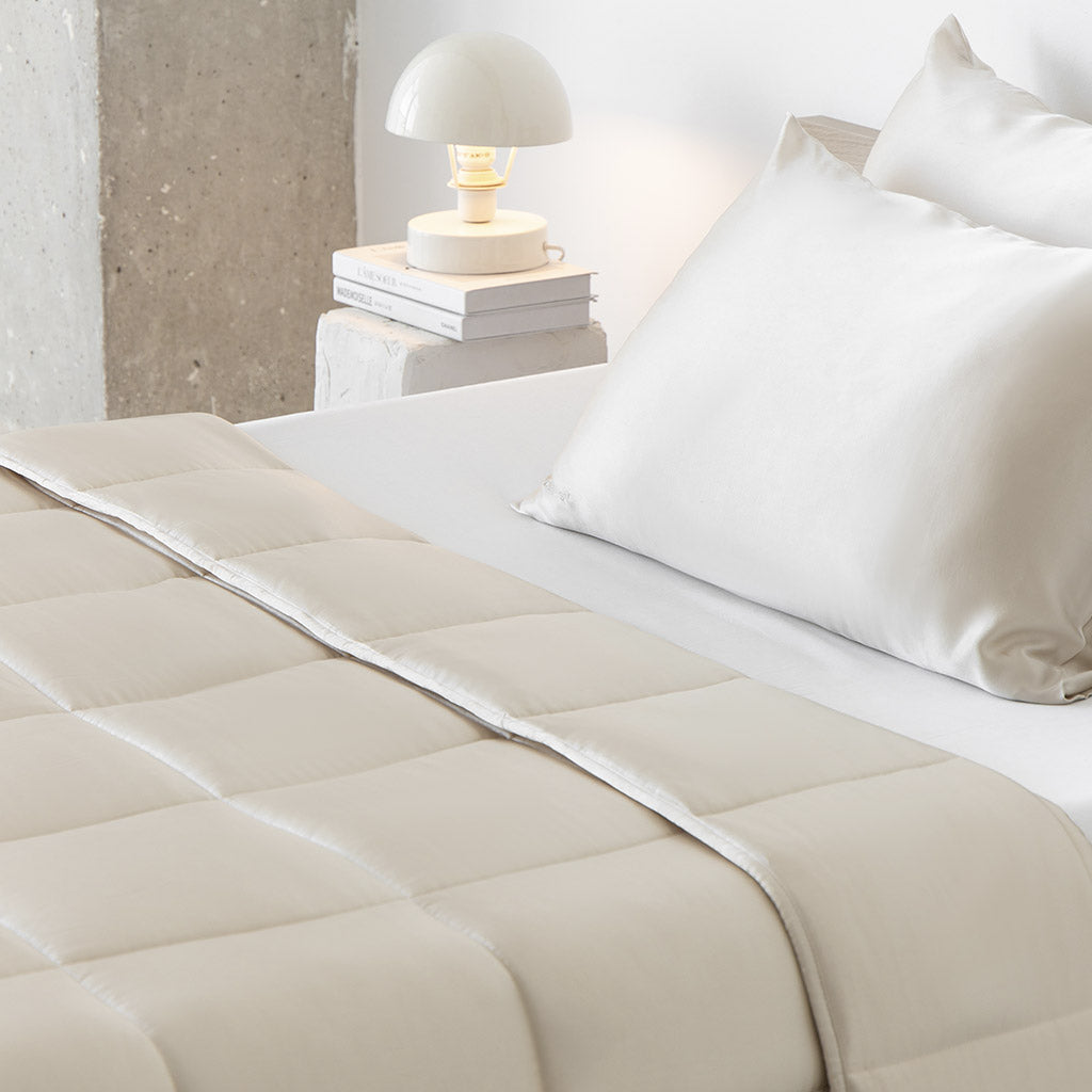 A neatly made bed with cream-colored sheets, pillows, and a Dusty Gold Silk Weighted Blanket creates a space for deep relaxation. A small lamp on stacked books rests on the nightstand, completing the modern minimalist look.