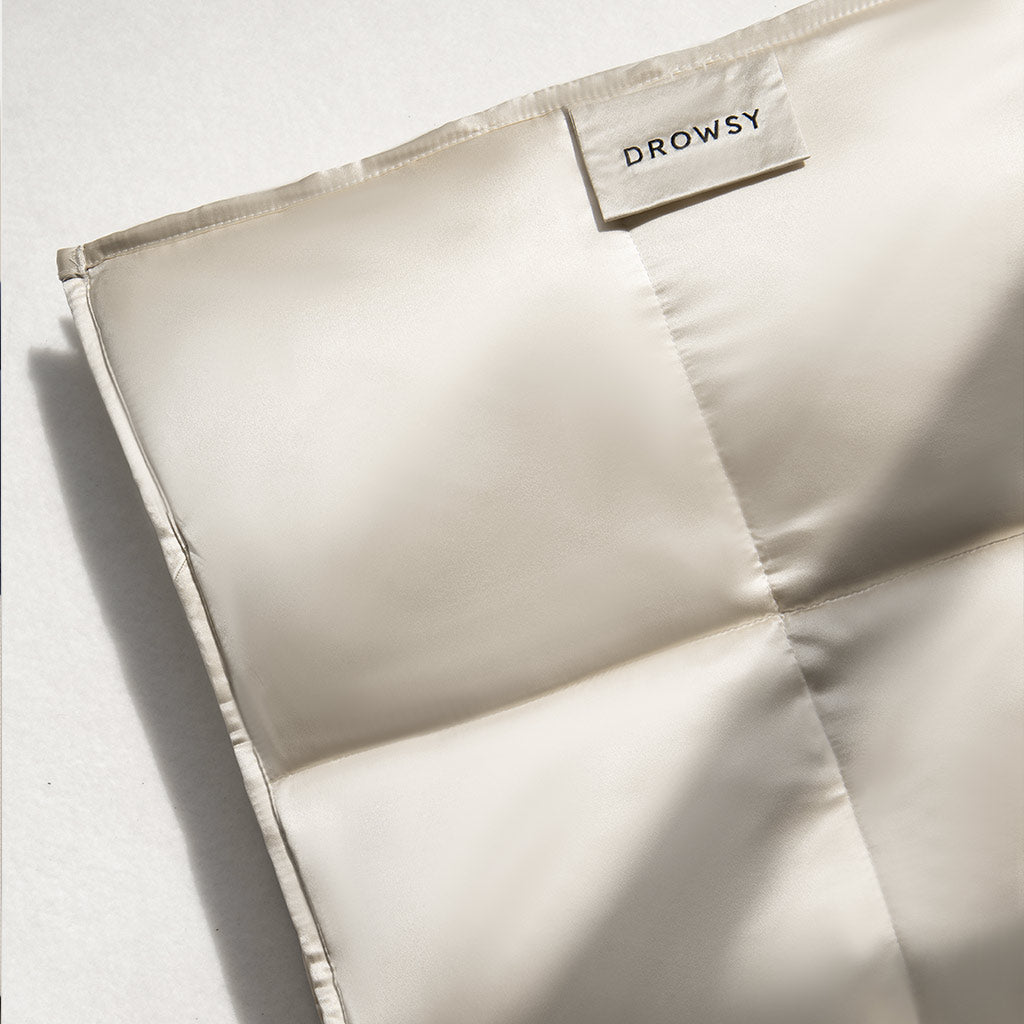 A close-up of the Dusty Gold Silk Weighted Blanket, showcasing its light beige, quilted mulberry silk with a satin finish. The surface looks smooth and soft, inviting deep relaxation.