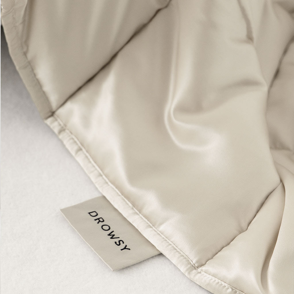 A close-up of the Dusty Gold Silk Weighted Blanket, featuring a visible DROWSY fabric tag, resting on a soft, light-colored surface—ideal for deep relaxation.