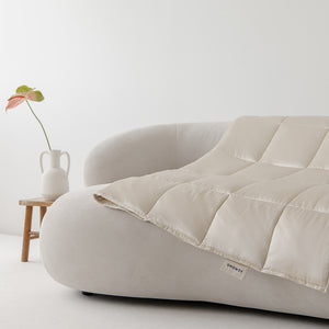 A Dusty Gold Silk Weighted Blanket draped over a curved off-white sofa, with a small wooden side table nearby holding a white ceramic vase and a single pink and green stem in the background.