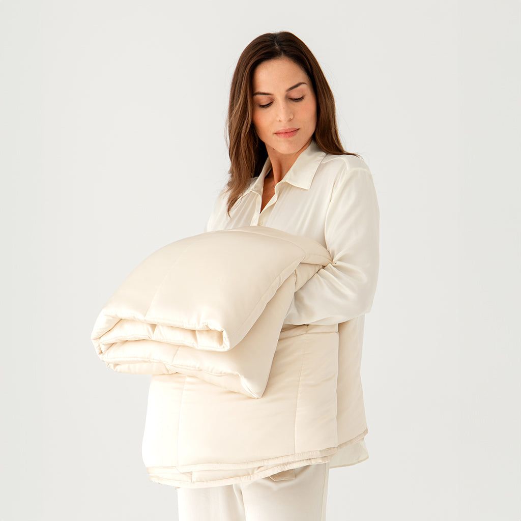 A woman in cream-colored pajamas holds a folded Dusty Gold Silk Weighted Blanket against a plain white background, evoking deep relaxation.