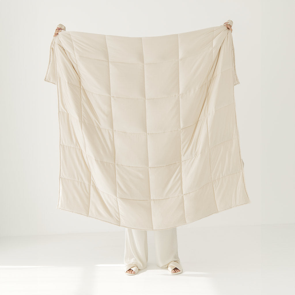 A person holds up the Dusty Gold Silk Weighted Blanket, a large beige quilted cover that hides most of their body except feet and fingertips, set against a plain white background for a calming, relaxed vibe.