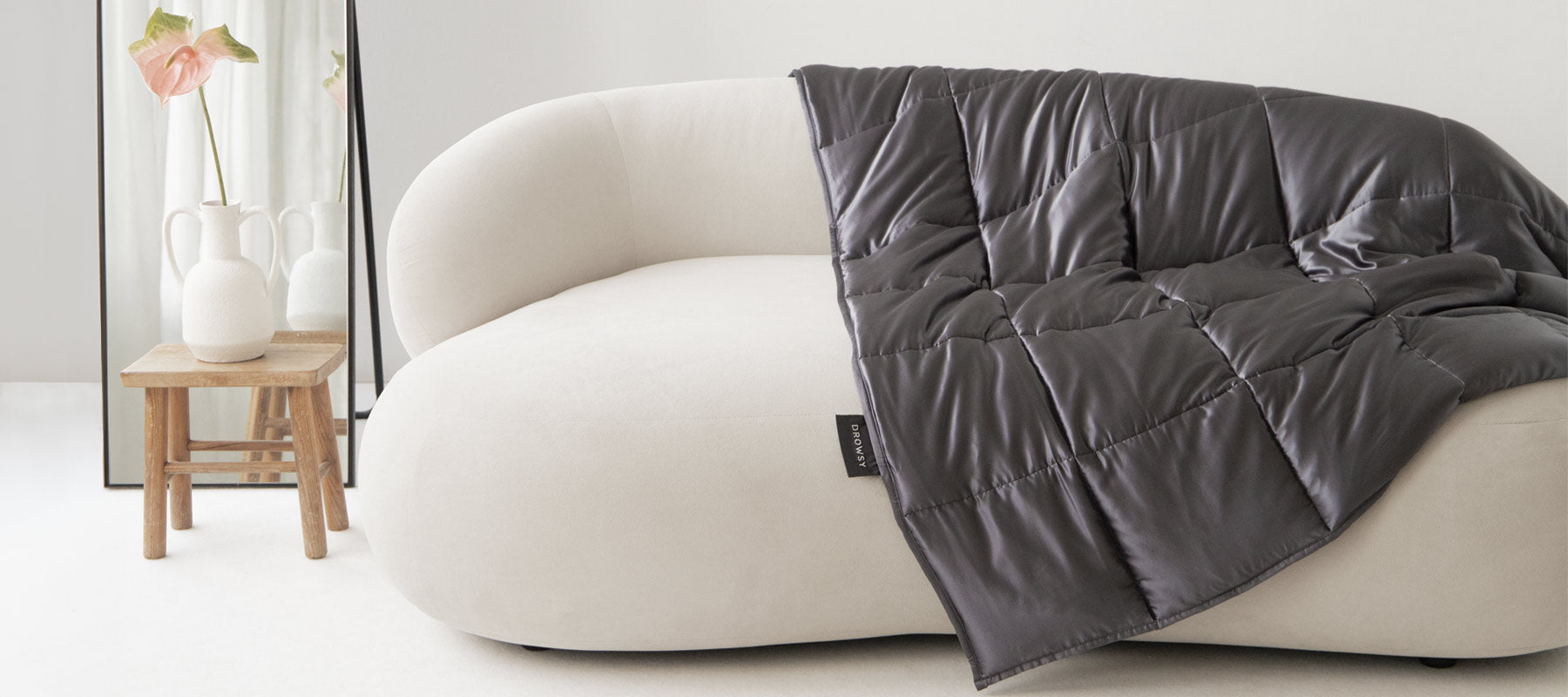 A modern white curved sofa with a dark quilted blanket draped over one side. Next to it, a small wooden stool holds two white vases and a tall pink flower. A large mirror leans against the wall in the background.