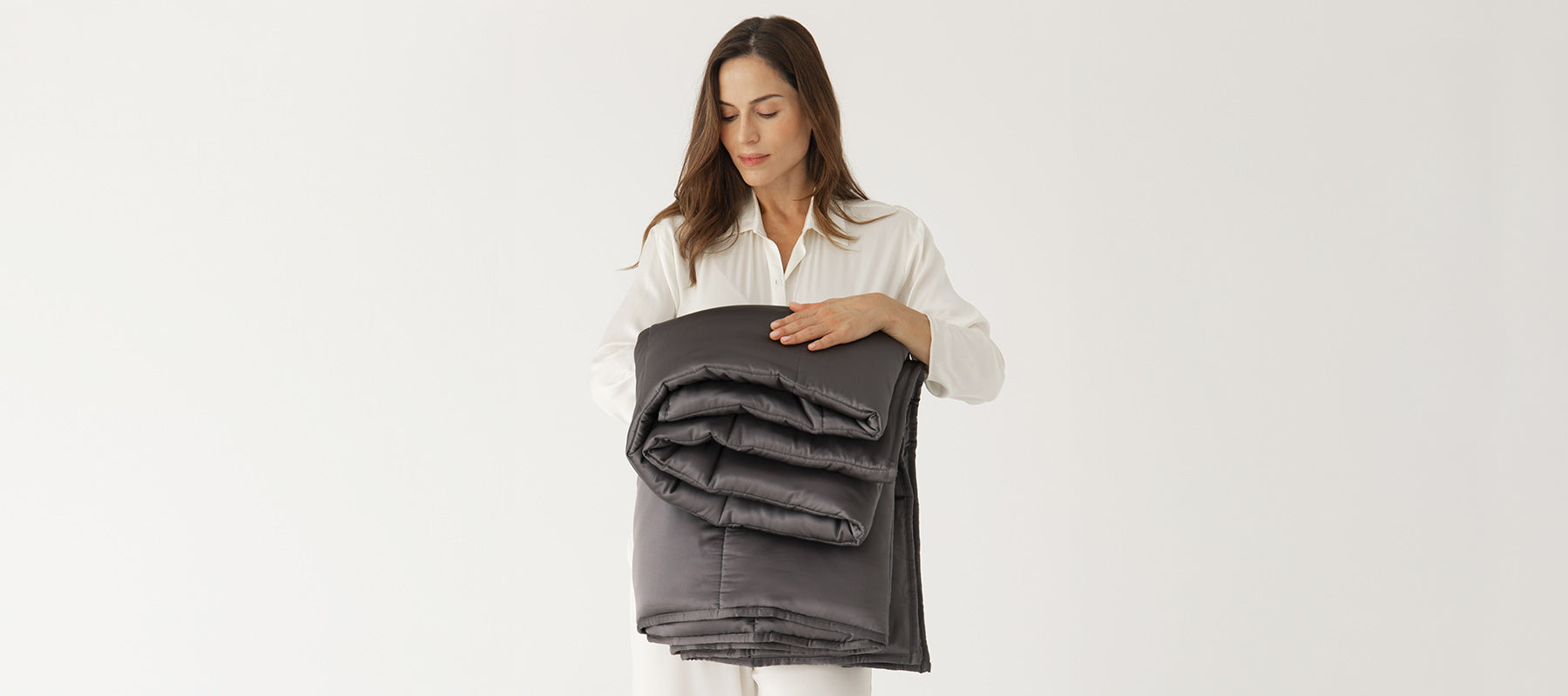 A woman wearing white pajamas stands against a plain white background, holding a neatly folded dark gray weighted blanket in her arms and looking down at it with a gentle expression.