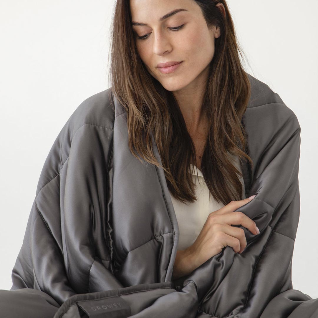 A woman with long brown hair drapes herself in the Moonlight Shadow Silk Weighted Blanket, its dark gray quilted silk wrapping her as she gazes down peacefully, embodying deep relaxation against a plain white background.