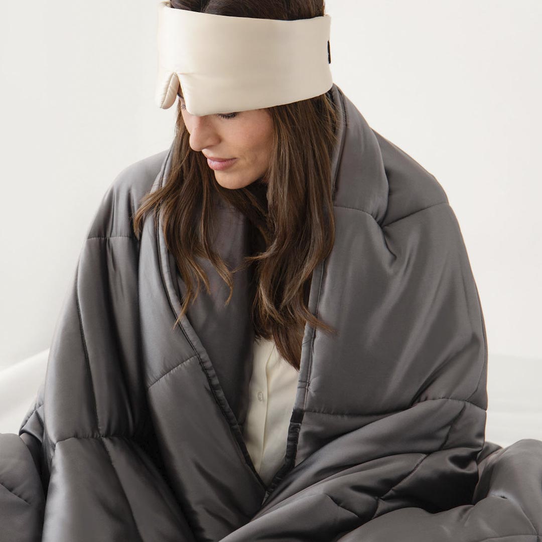 A woman wearing a cream-colored sleep mask sits wrapped in a large, gray comforter, looking down with a calm expression. The background is plain and light-colored.