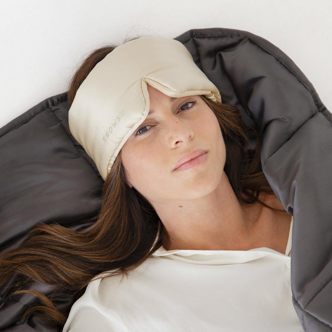 A woman in a white top lies on a soft surface, draped in the Moonlight Shadow Silk Weighted Blanket and wearing a light silk eye mask, showcasing ultimate relaxation and comfort.