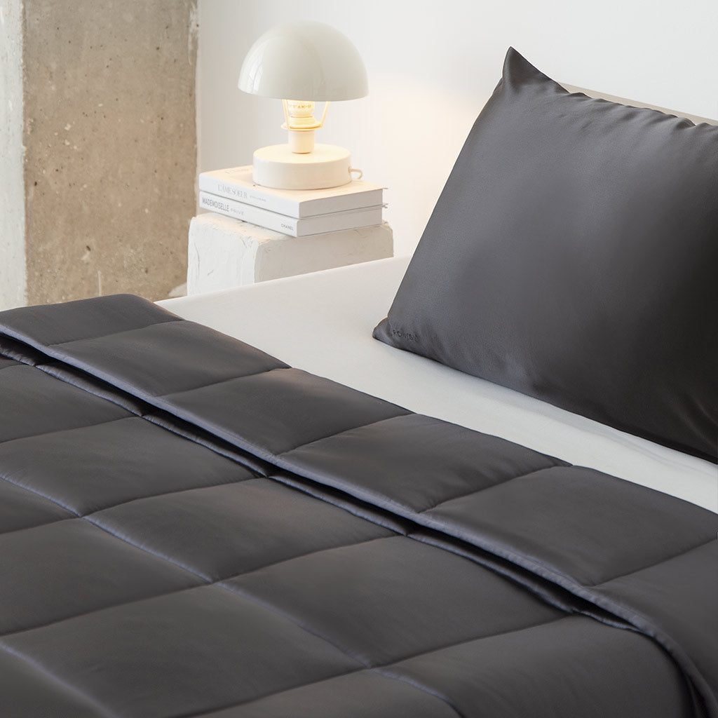 A neatly made bed with dark gray bedding and pillow sits by a white nightstand holding stacked books and a small round lamp, creating a modern, minimalist space perfect for unwinding with the Moonlight Shadow Silk Weighted Blanket.