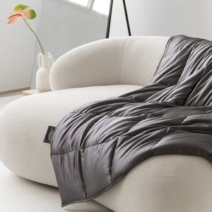 The Moonlight Shadow Silk Weighted Blanket in dark gray is draped over a modern white curved sofa in a minimalist room, with a vase and single plant in the background, creating an inviting space for deep relaxation.
