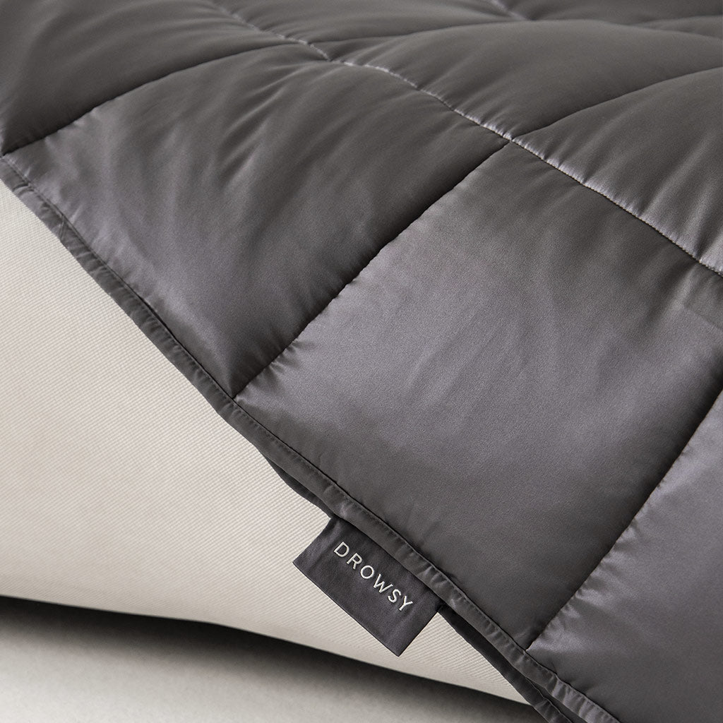 A close-up of the Moonlight Shadow Silk Weighted Blanket, dark gray and quilted with a “DROWSY” label, draped over a light beige surface. Its stitching and smooth mulberry silk texture suggest deep relaxation.