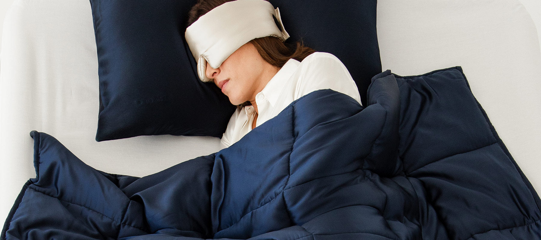 A person wearing a white sleep mask is lying on their side in bed, covered with a dark blue comforter and resting their head on a matching pillow.