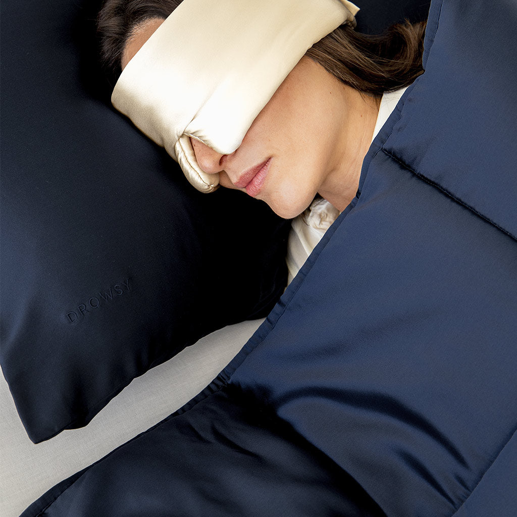 A person wearing a silk sleep mask rests on a dark blue satin pillow and is covered with a matching blue blanket. The word DROWSY is embroidered on the pillow.
