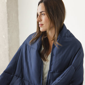 A woman with straight brown hair looks to the side indoors, draped in a Midnight Blue Silk Weighted Blanket. A light-colored wall is in the background as she enjoys deep relaxation.
