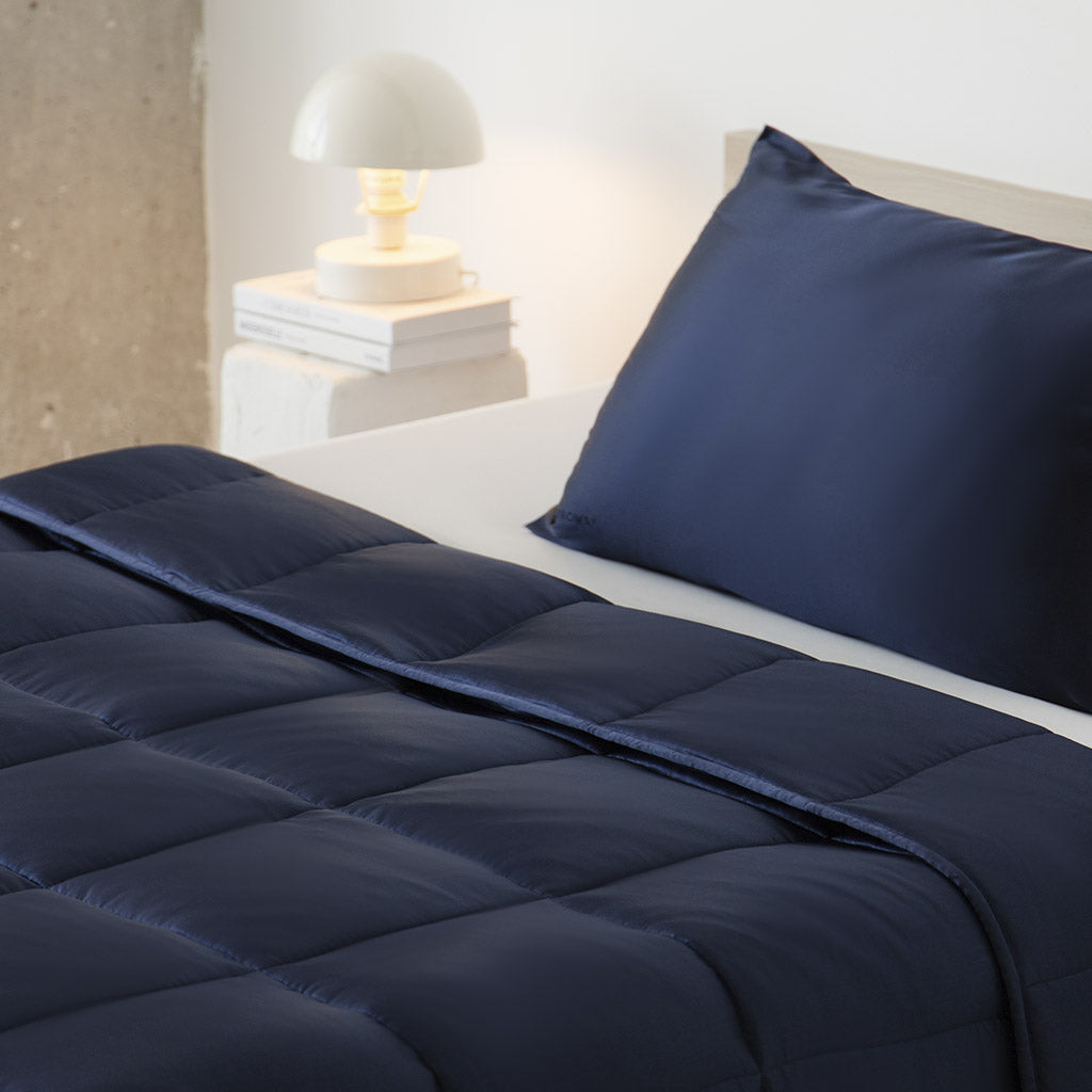 A made bed with a navy blue comforter and matching pillowcase features a Midnight Blue Silk Weighted Blanket for deep relaxation. A white lamp and stacked books sit on the bedside table next to a light-colored wall and concrete pillar.